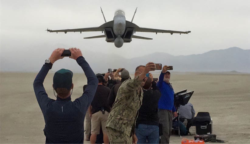 A group of people are standing on a beach, watching a military jet flying low overhead. Some of the people are taking pictures of the jet with their cell phones. AI generated content