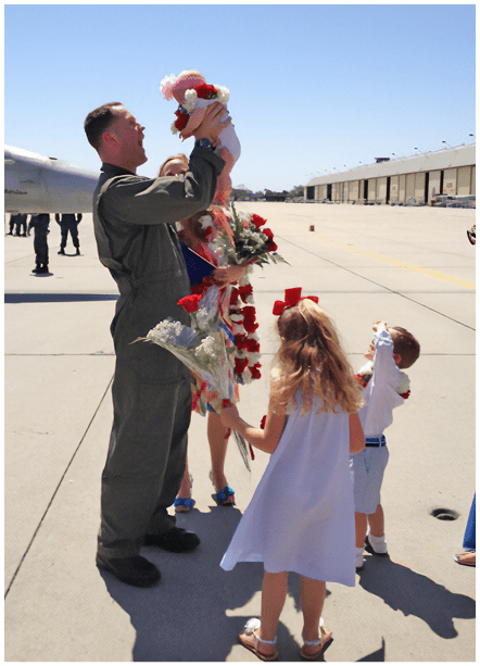 A man in military uniform holds a baby while a woman in a white dress holds flowers. They are surrounded by other people. AI generated content